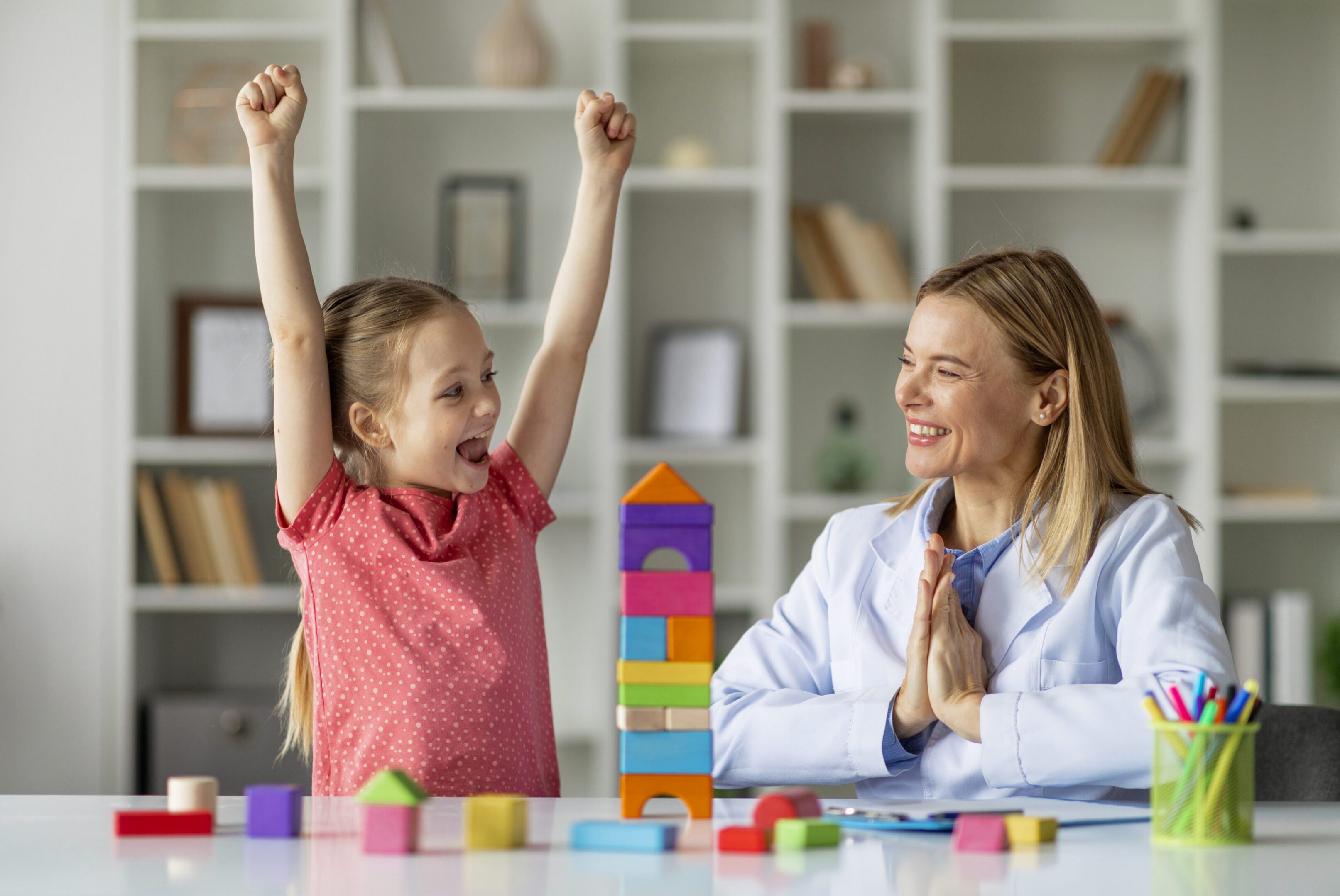 Cute Little Girl And Child Development Specialist Celebrating Therapy Progress During Meeting In Office, Smiling Therapist Lady Clapping Hands While Happy Female Kid Raising Fists And Shouting Hooray
