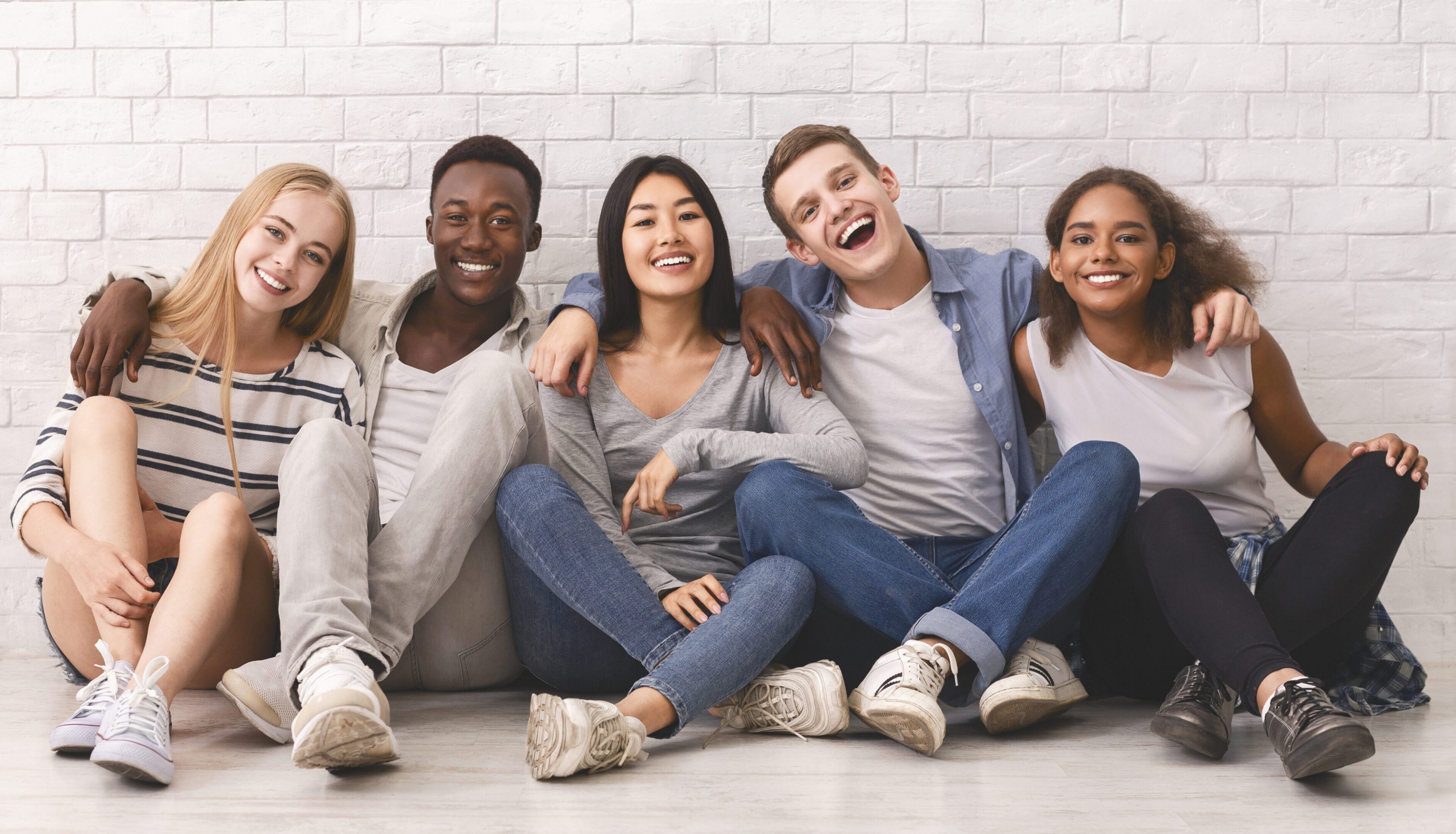 Multiracial students sitting on floor, embracing and having fun over white brick wall, posing on camera